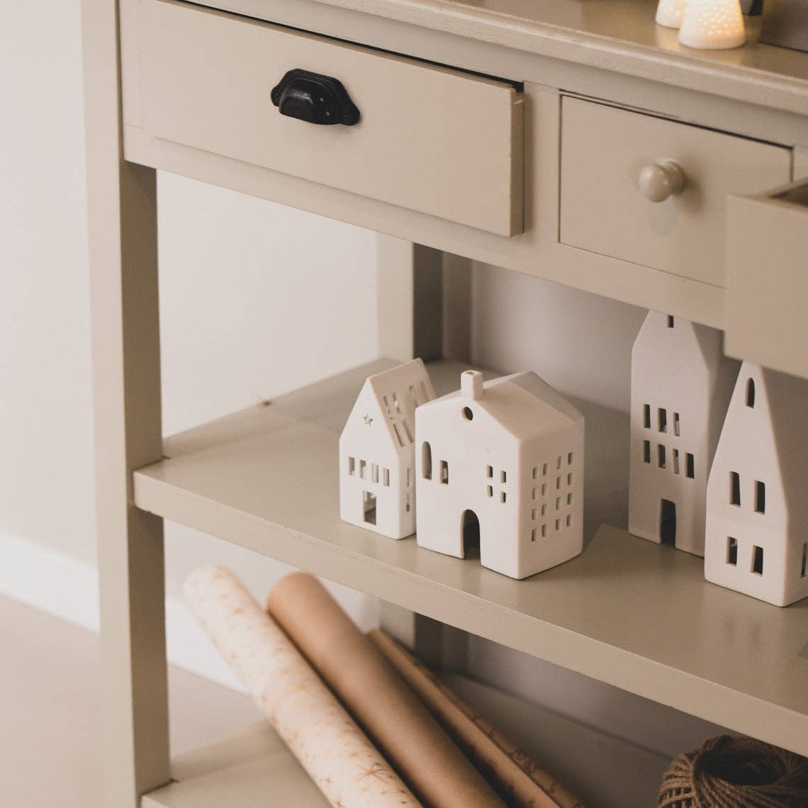 Small white porcelain houses on a shelf next to a wooden cabinet with drawers.