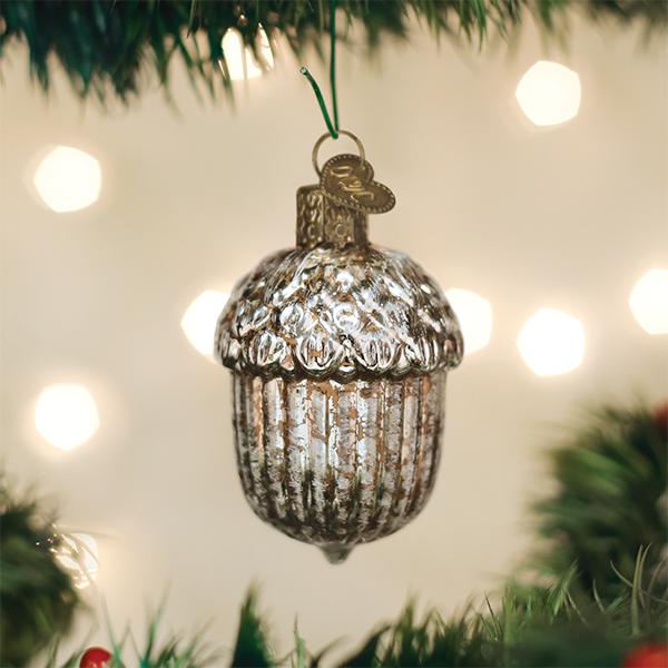 A silver acorn-shaped Christmas ornament with a textured, ribbed body and a small hanging loop, shown hanging from a green pine branch against a warm, blurred background with bokeh lights.