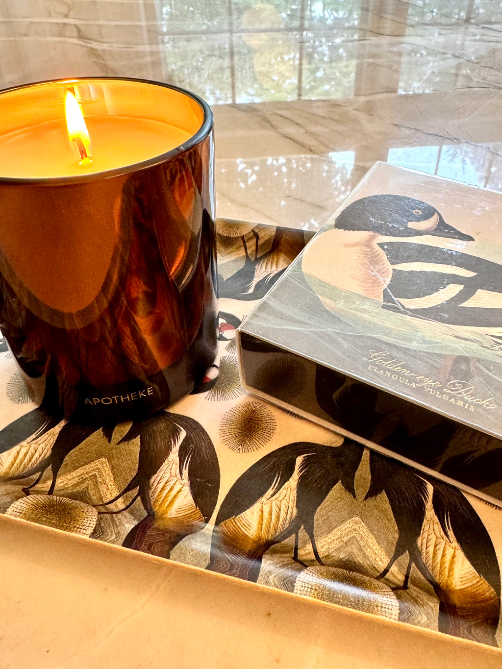 A lit amber glass Apotheke Pumpkin Ginger candle and a decorative Archivist duck-themed box resting on a patterned tray in a real indoor setting with a window and marble-like surface visible in the background.
