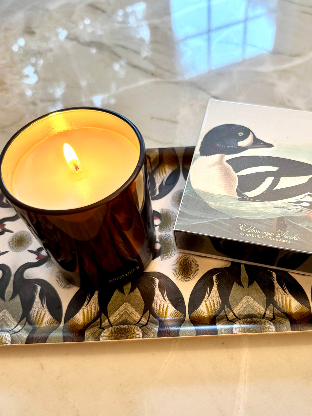 A lit amber glass candle with a warm yellow flame sits on a patterned tray next to its illustrated product box featuring a duck illustration.