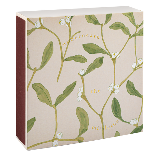 An Archivist square match box with a mistletoe botanical print featuring green leaves and white berries on a pale blush-beige background, with the words 'underneath the mistletoe' printed in gold. The side of the box is a solid dark red-brown color.