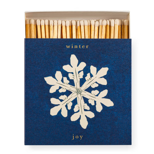 An Archivist square matchbox with a textured dark blue cover featuring a large white snowflake and the words 'winter' and 'joy' in gold. The top of the box is open, showing a row of natural wood matches inside, on a plain white background.