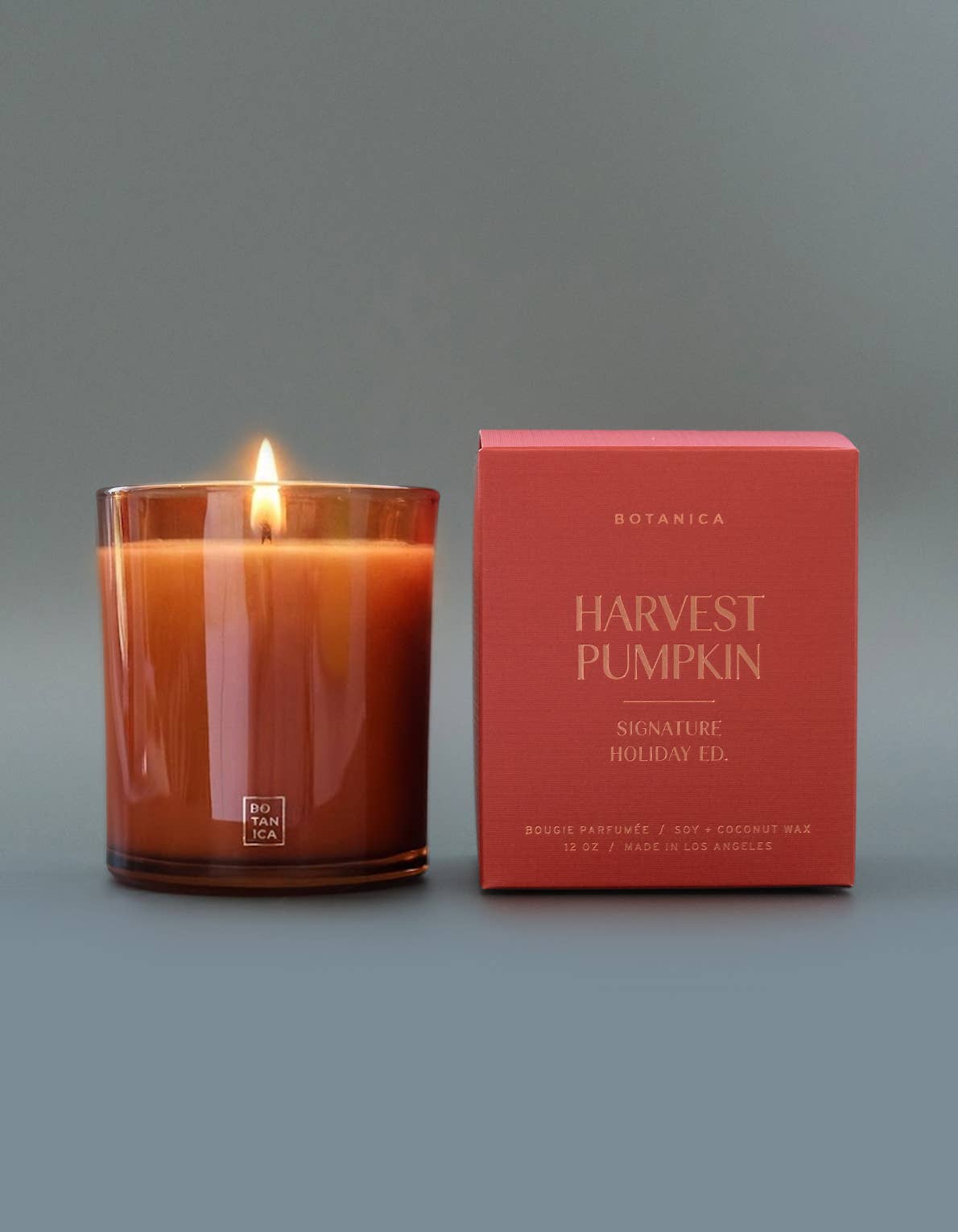 A lit Botanica 'Harvest Pumpkin' scented candle in a clear amber glass jar sits next to its orange packaging box on a neutral gray background.
