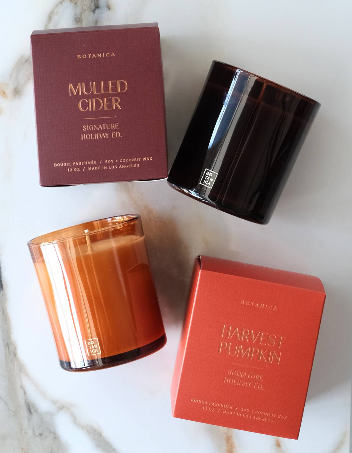 A Botanica Mulled Cider candle with its burgundy packaging next to a Botanica Harvest Pumpkin Candle with its orange packaging on a marble surface