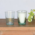 Two green glass votive holders with a candle and a flower on a wooden surface