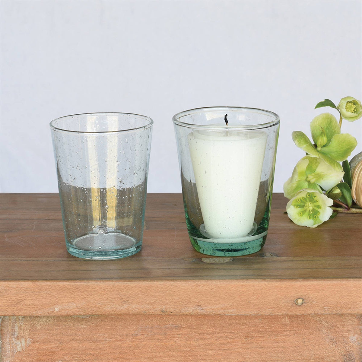 Two green glass votive holders with a candle and a flower on a wooden surface