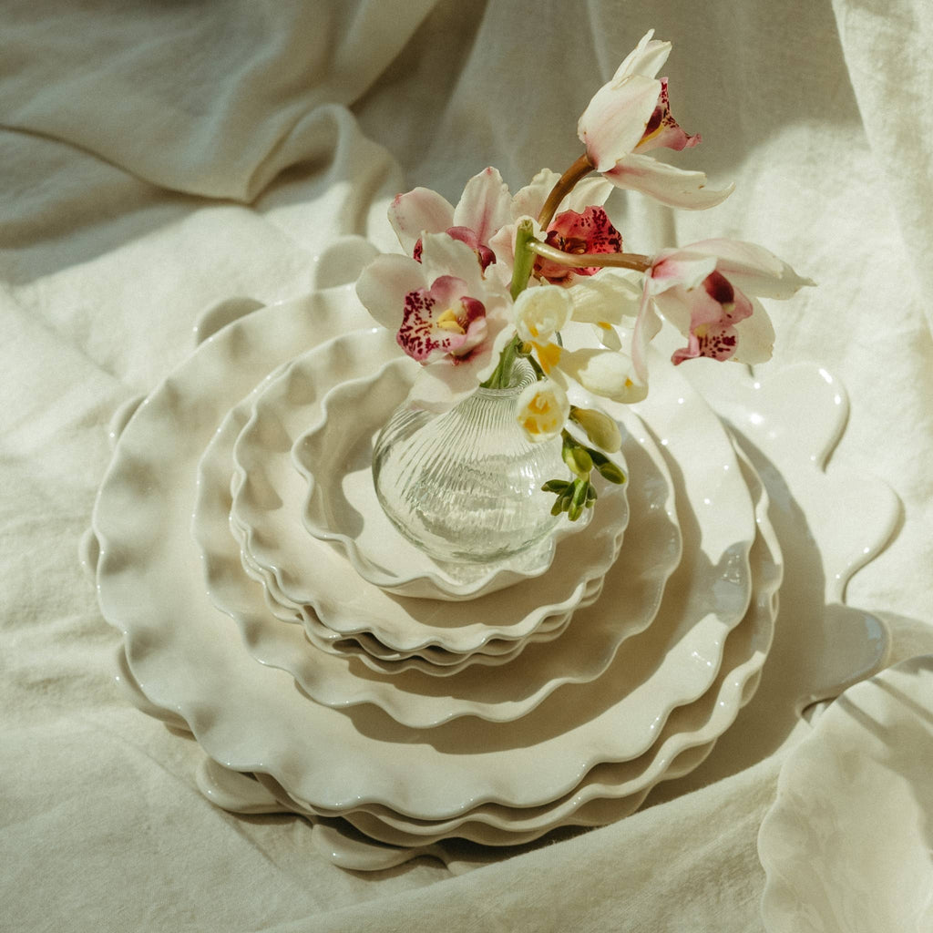 Stack of white ceramic Calypso plates with a ruffled edge with a small vase and flowers on a textured beige surface