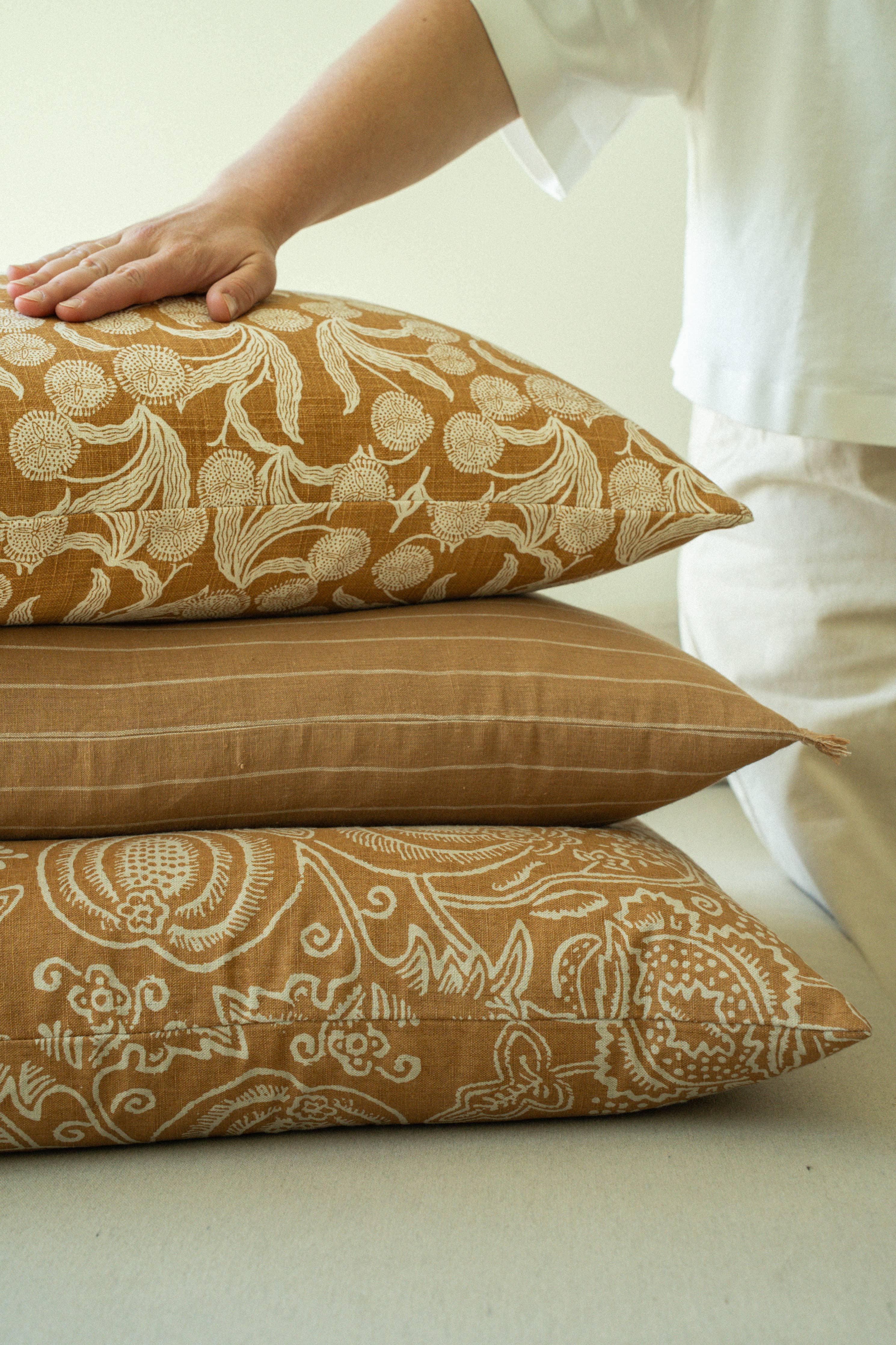 Three patterned pillows on a light surface with a person's hand and leg in the background.