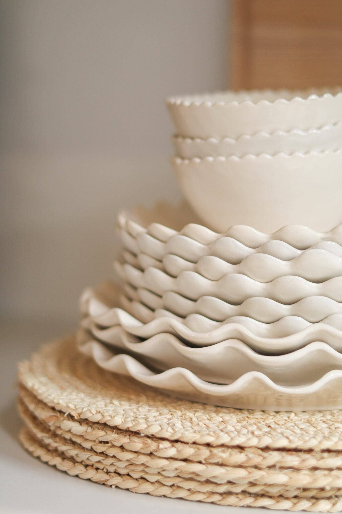 A stack of cream-colored ceramic Calypso tableware, including scalloped plates and a bowl, resting on woven natural fiber placemats against a softly blurred neutral background.