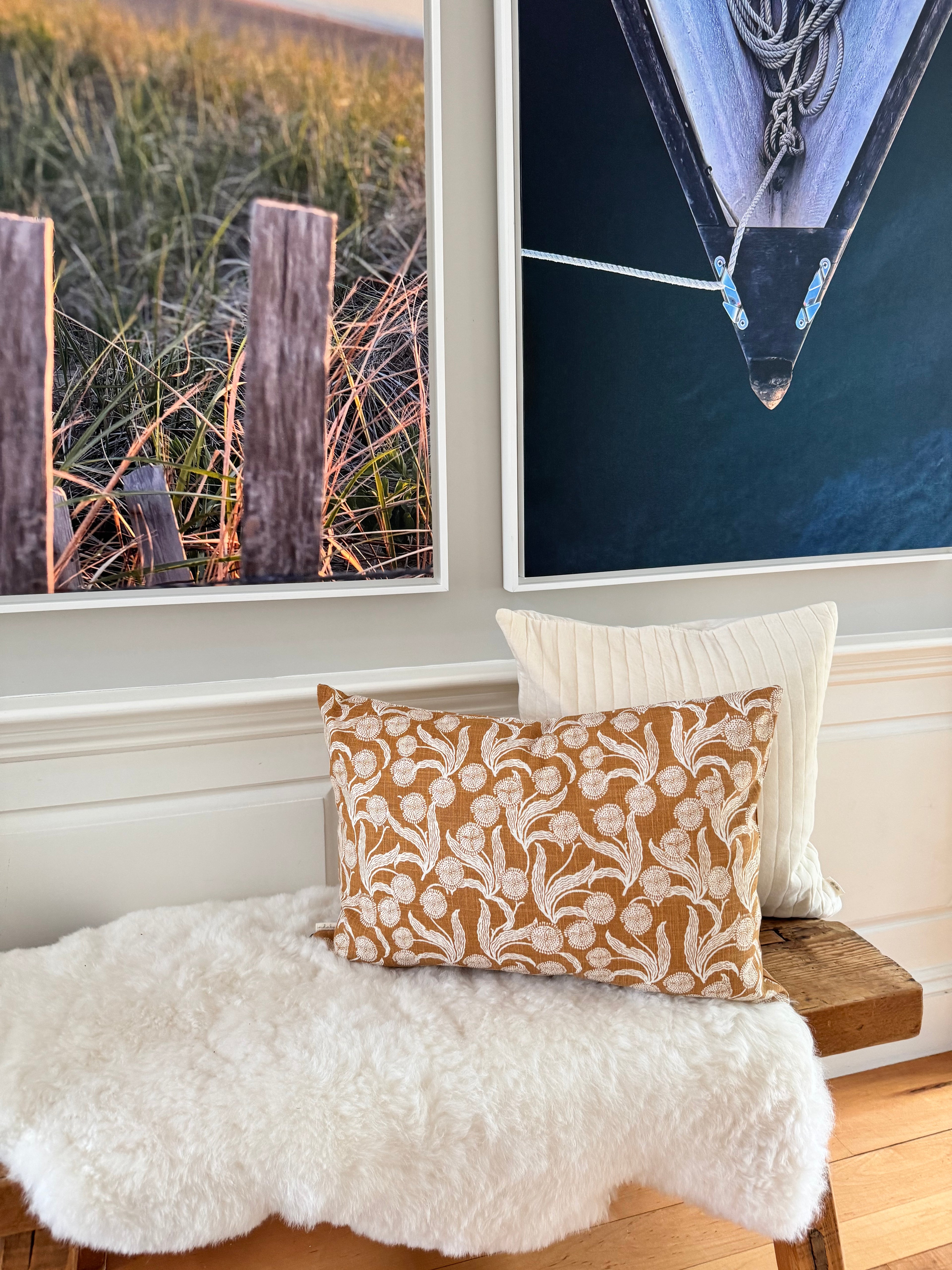 A styled interior scene with two framed wall art prints above a wooden bench draped in white faux fur, with two decorative pillows in front: one tan and cream floral patterned and one solid cream.