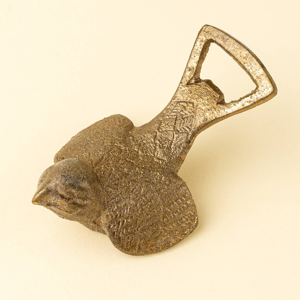 A rustic cast iron bird-shaped bottle opener shaped like a sparrow, featuring a textured surface and a functional D-shaped opener tab, set against a solid cream background. Brass finish