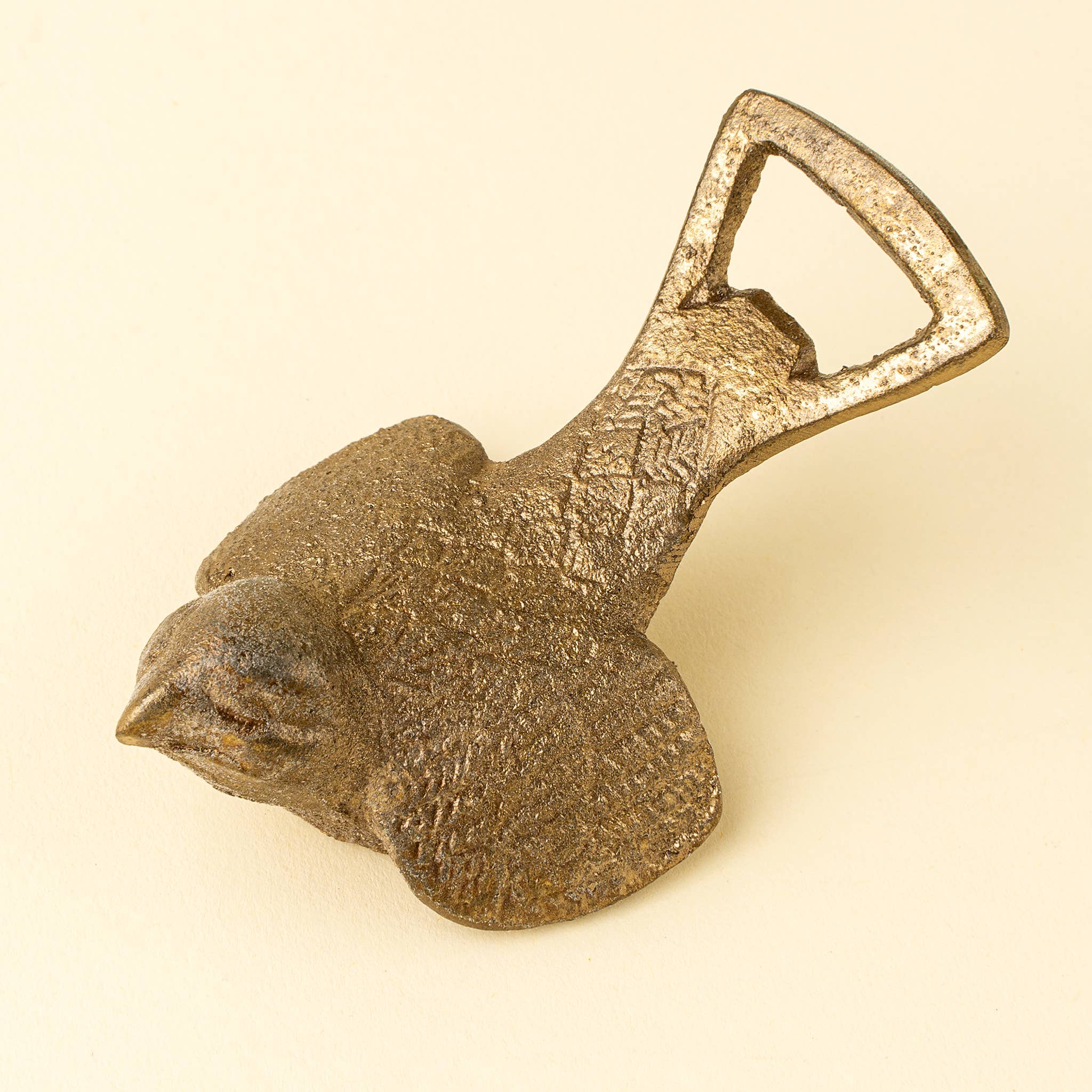 A rustic cast iron bird-shaped bottle opener shaped like a sparrow, featuring a textured surface and a functional D-shaped opener tab, set against a solid cream background. Brass finish