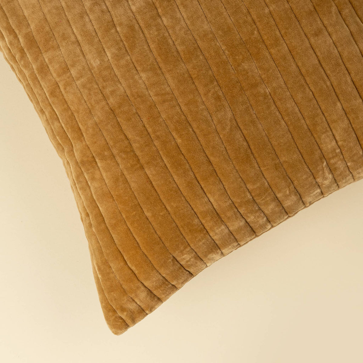 A close-up of a square throw pillow in a warm camel brown velvet fabric, showing the vertical ribbed texture against a solid cream background.