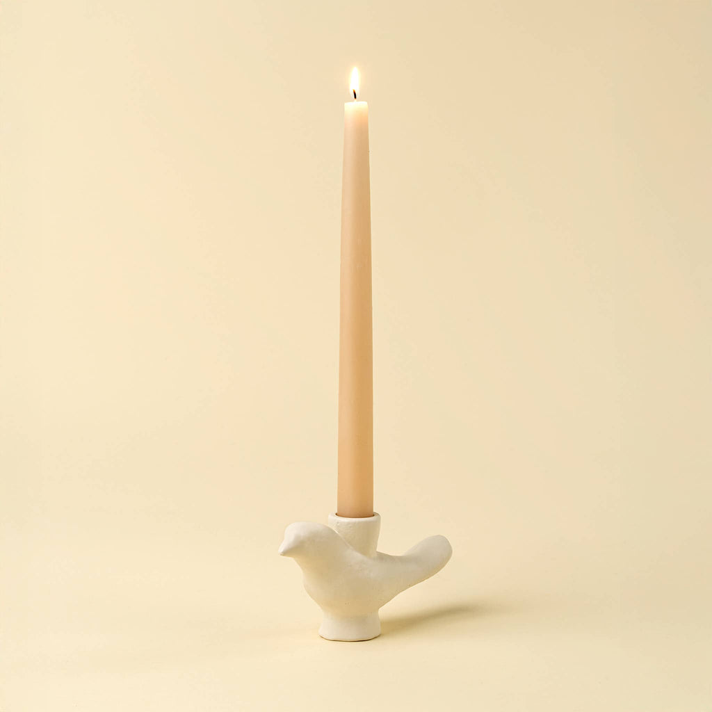A lit taper candle in a white bird-shaped holder against a warm cream background.