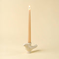 A lit taper candle in a white bird-shaped holder against a warm cream background.