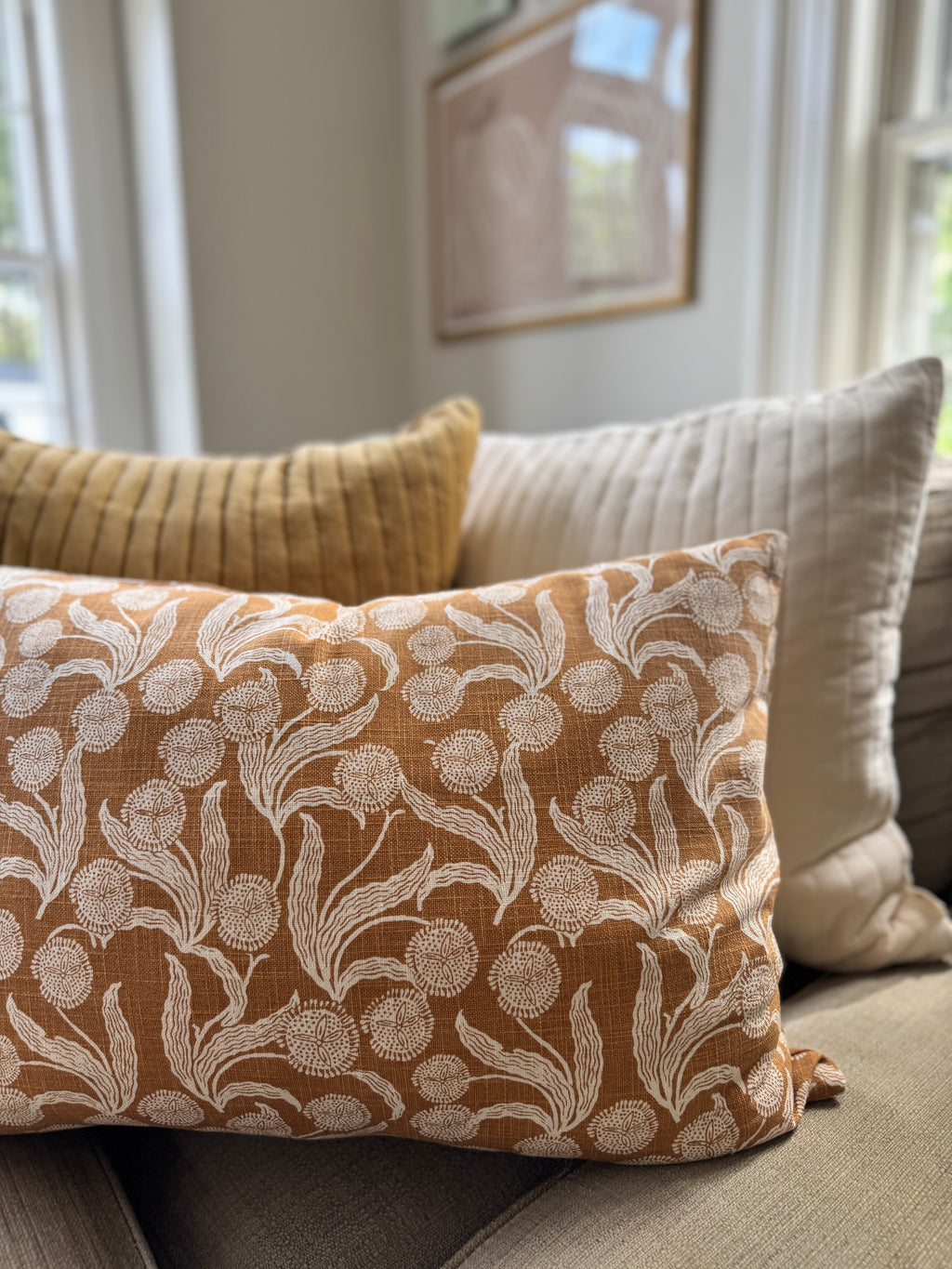 A rectangular throw pillow with a cream floral block-print pattern on a warm ochre-brown linen fabric, styled on a sofa with other decorative pillows in a bright living room setting.