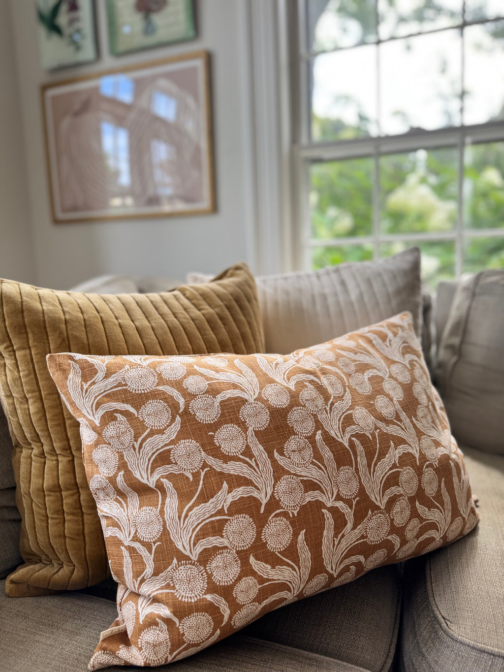 A rectangular decorative throw pillow with a cream floral damask pattern on a warm rust-orange background, styled on a gray sofa next to a mustard yellow quilted pillow in a bright living room.