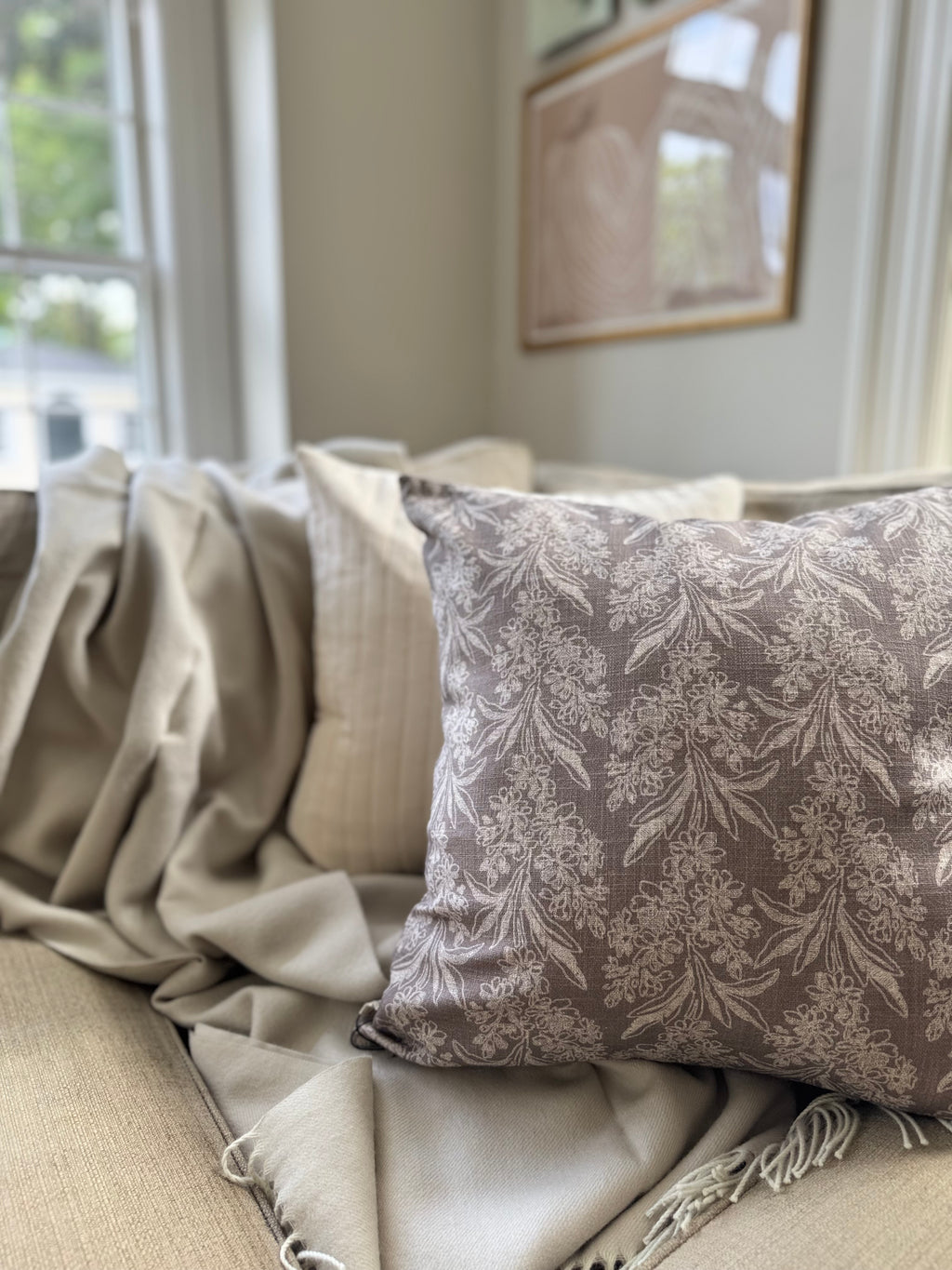 A decorative square throw pillow with a blue gray damask floral pattern on a taupe background, styled on a beige sofa with a cream throw blanket.