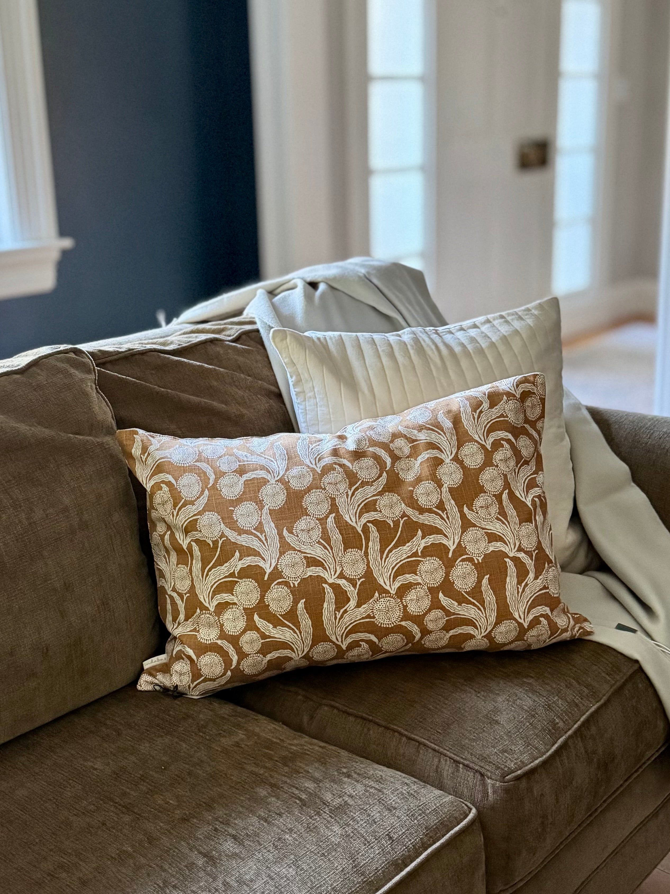 A decorative throw pillow with a cream floral damask pattern on a warm brown background, styled on a brown fabric sofa next to a white pillow and a cream throw blanket.