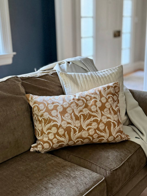 A decorative throw pillow with a cream floral damask pattern on a warm brown background, styled on a brown fabric sofa next to a white pillow and a cream throw blanket.