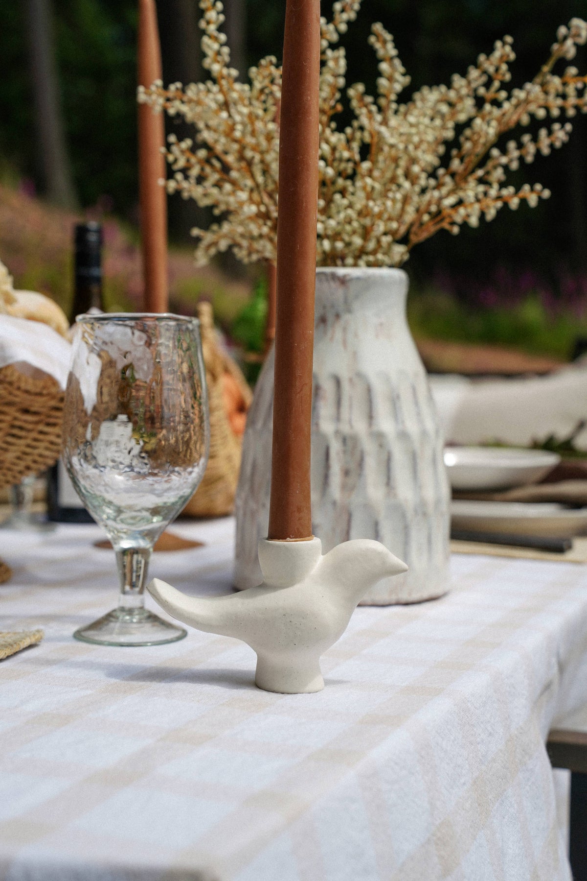A white ceramic bird-shaped candlestick holder with a tall brown taper candle, set on a white checkered tablecloth next to a clear textured wine glass, with a textured white vase of dried flowers in the background.