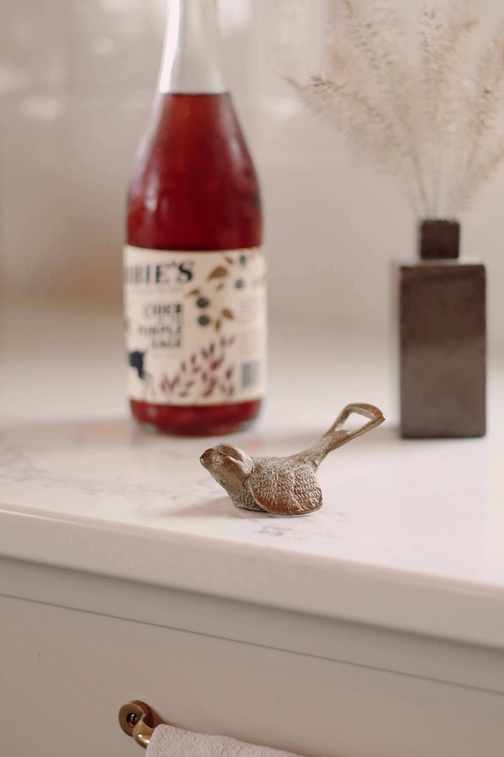 A bottle with a decorative bird-shaped bottle opener resting in front of it on a white marble countertop, with a vase of dried grass in the background.