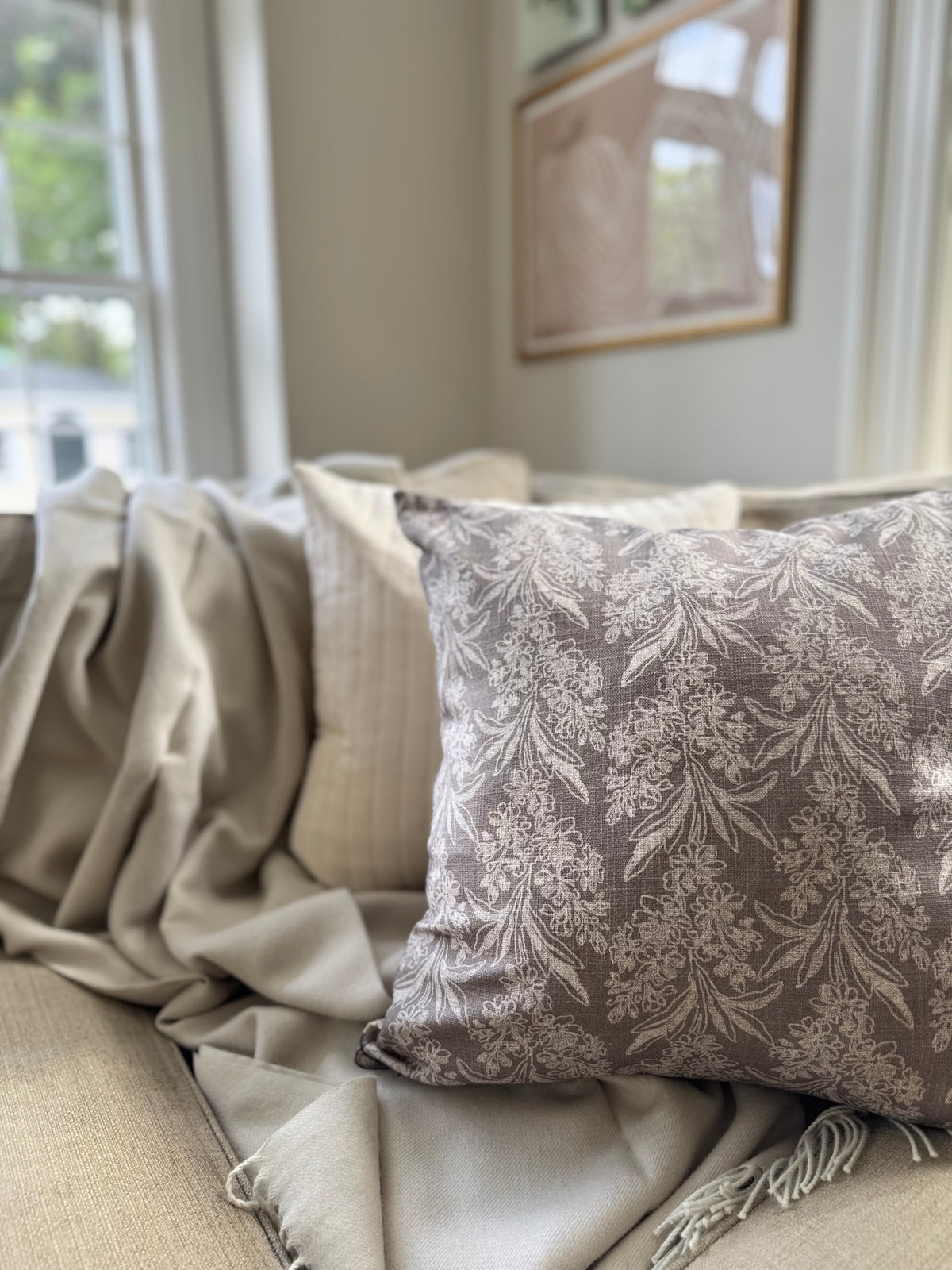 A square decorative throw Indaba pillow with a beige damask floral pattern on a taupe-gray background, styled with a cream velvet Indaba square throw pillow on a beige sofa with a hazelnut shupaca alpaca throw blanket in a softly lit living room.