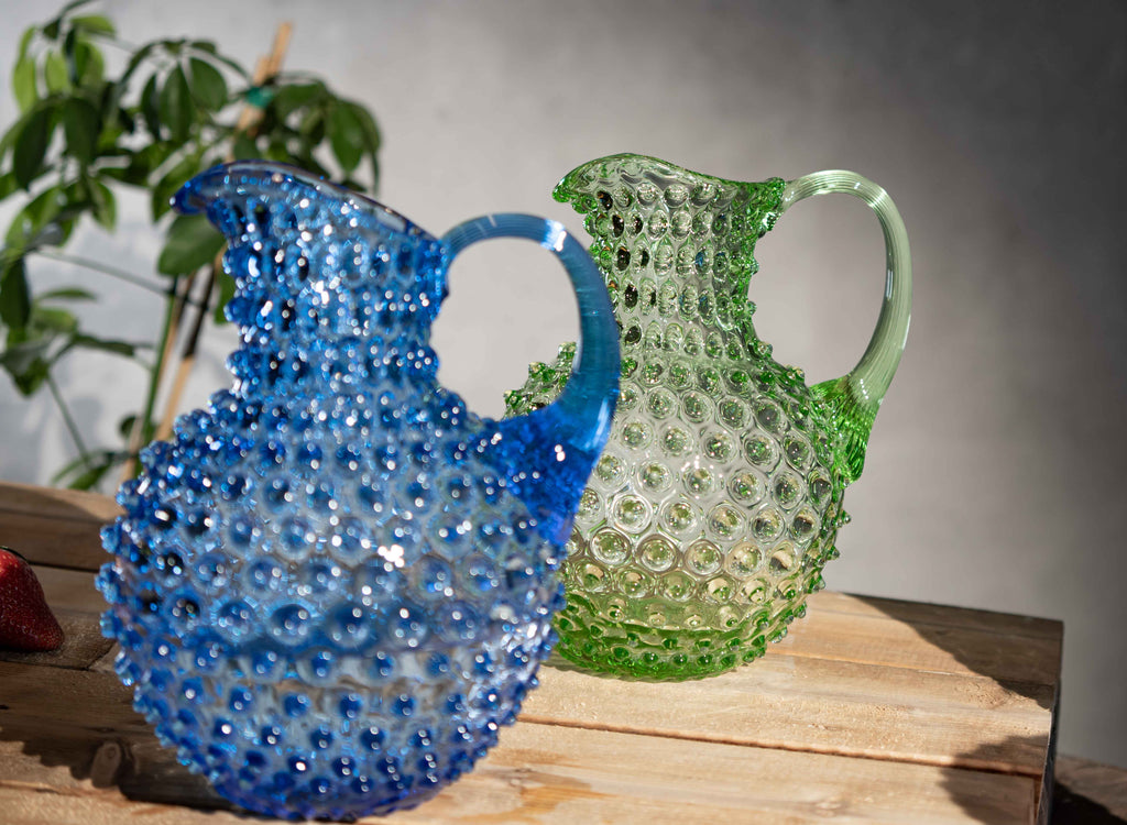 Blue and green Klimchi hobnail glass pitchers on a wooden surface with a plant in the background.