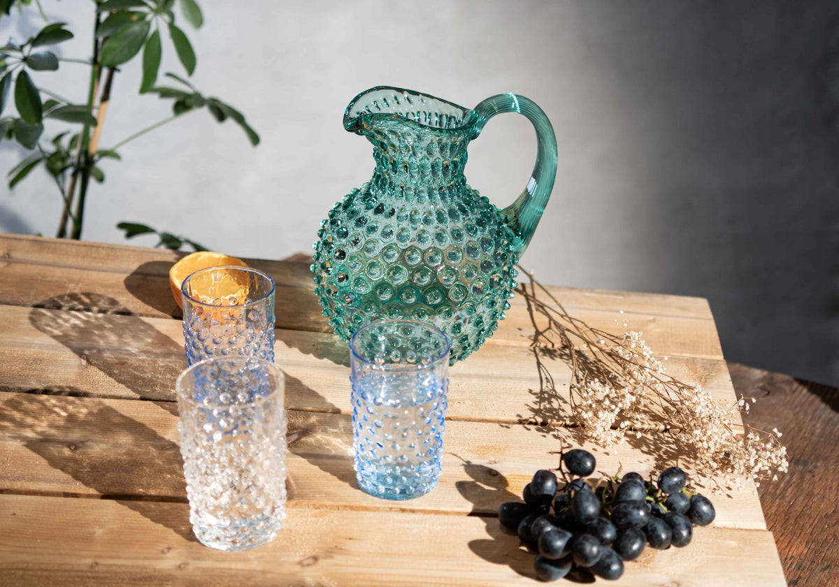 Teal glass Klimchi hobnail pitcher and clear glasses on a wooden surface with grapes and dried plants.