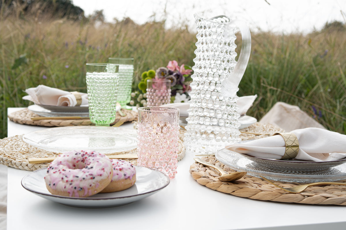 Outdoor table setting with donuts, Klimchi hobnail tumblers in pink and green, a Klimchi hobnail tall glass jug and cutlery in a natural setting