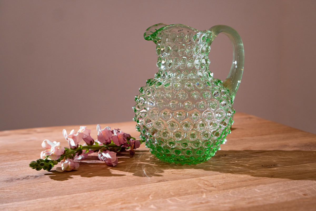 Green Klimchi hobnail glass pitcher on a wooden surface with pink flowers
