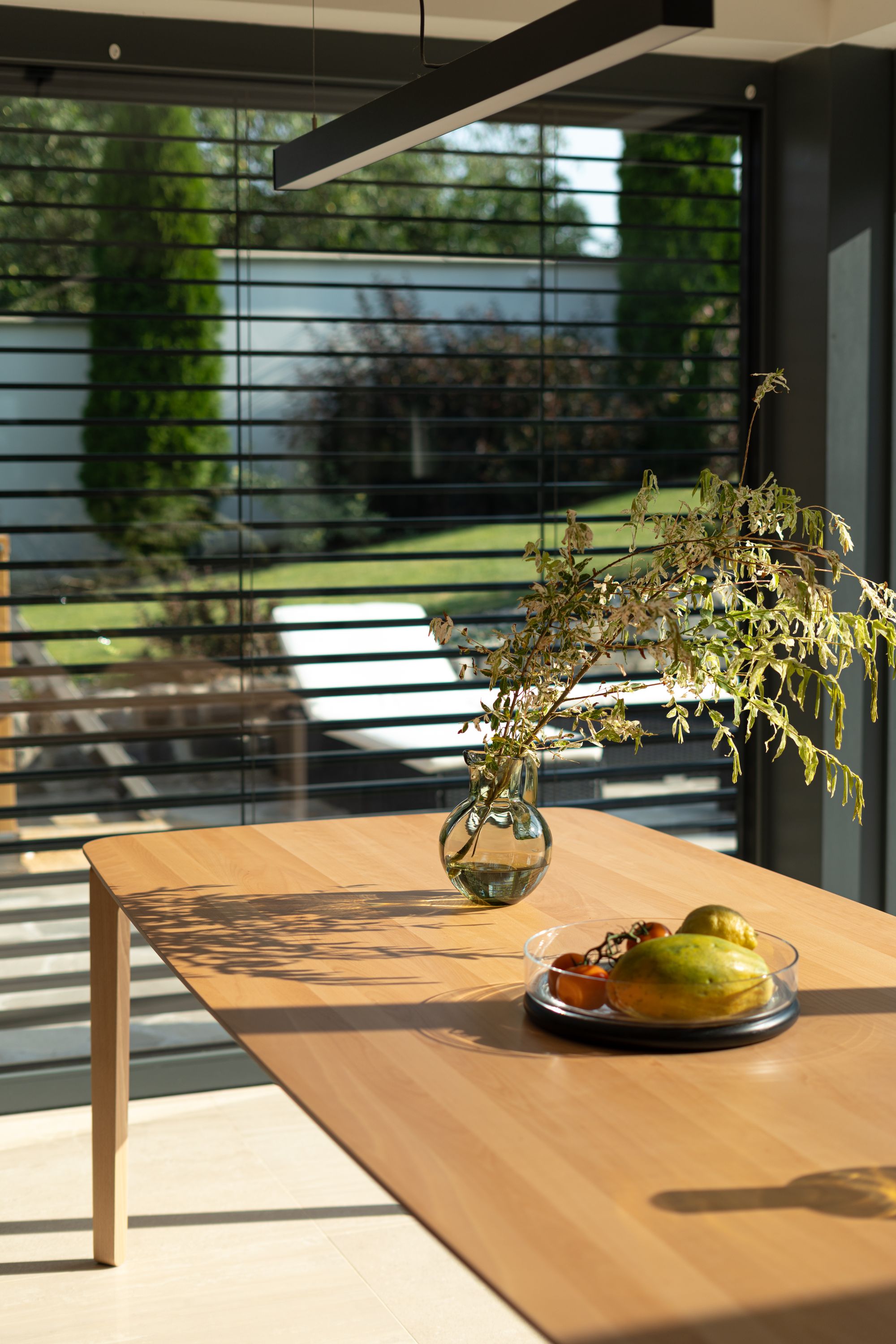 Wooden table with a Klimchi Marika glass jug and fruit plate in a modern indoor setting with large windows.