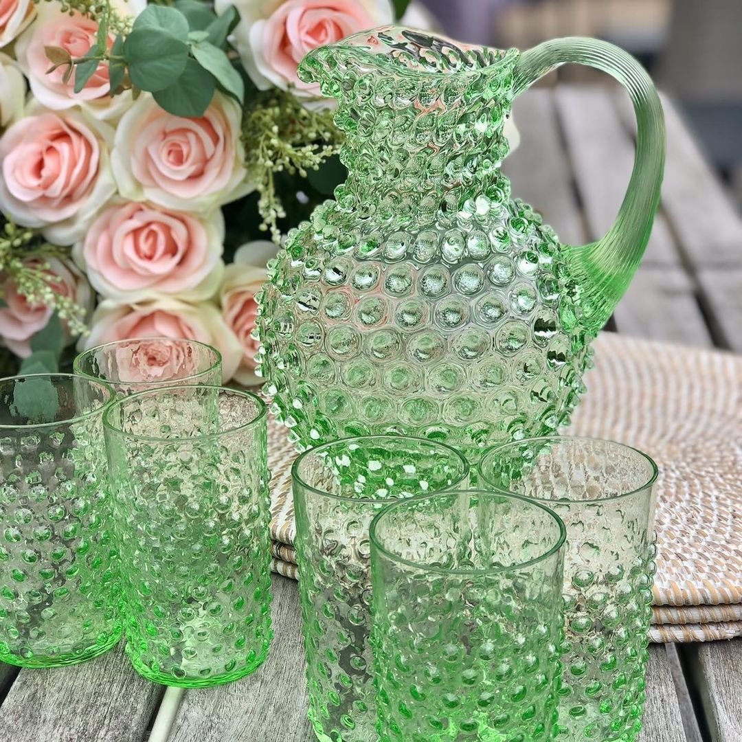 Green glass Klimchi hobnail pitcher and tumblers on a wooden surface with flowers in the background