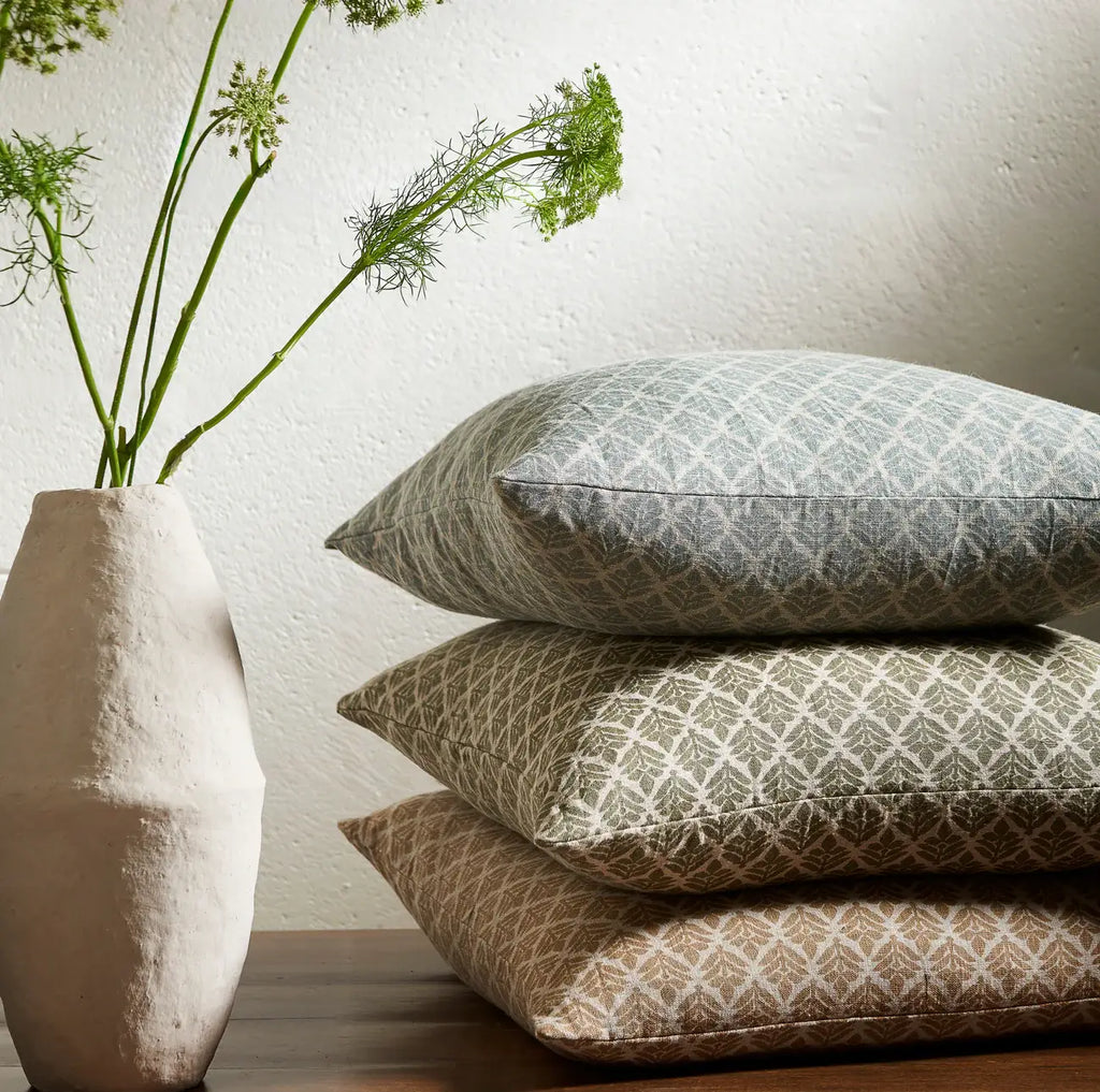Stack of Liam and Lana floral patterned linen pillows with down fill in 3 different colors with a vase containing greenery on a neutral background