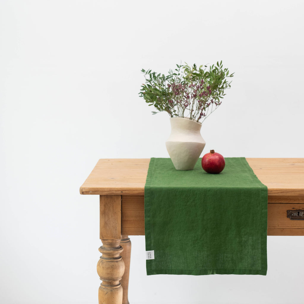 Green linen table runner on a wooden table with a vase and pomegranate.