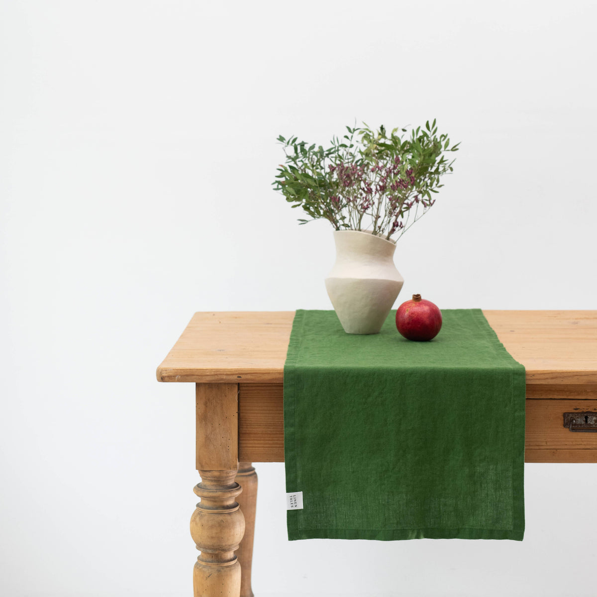 Green linen table runner on a wooden table with a vase and pomegranate.