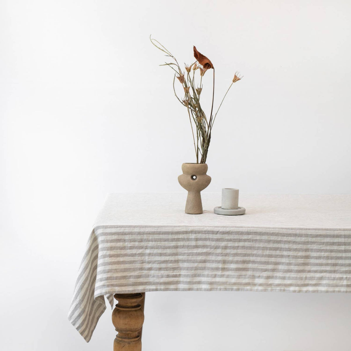 Table with a white and taupe striped linen tablecloth, vase with dried flowers, and candle on a white background