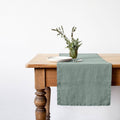 Milieu green linen table runner on a wooden table with a white plate and vase of greenery.