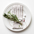 White plate with a white and natural striped linen napkin, greenery, and white balls on a white background