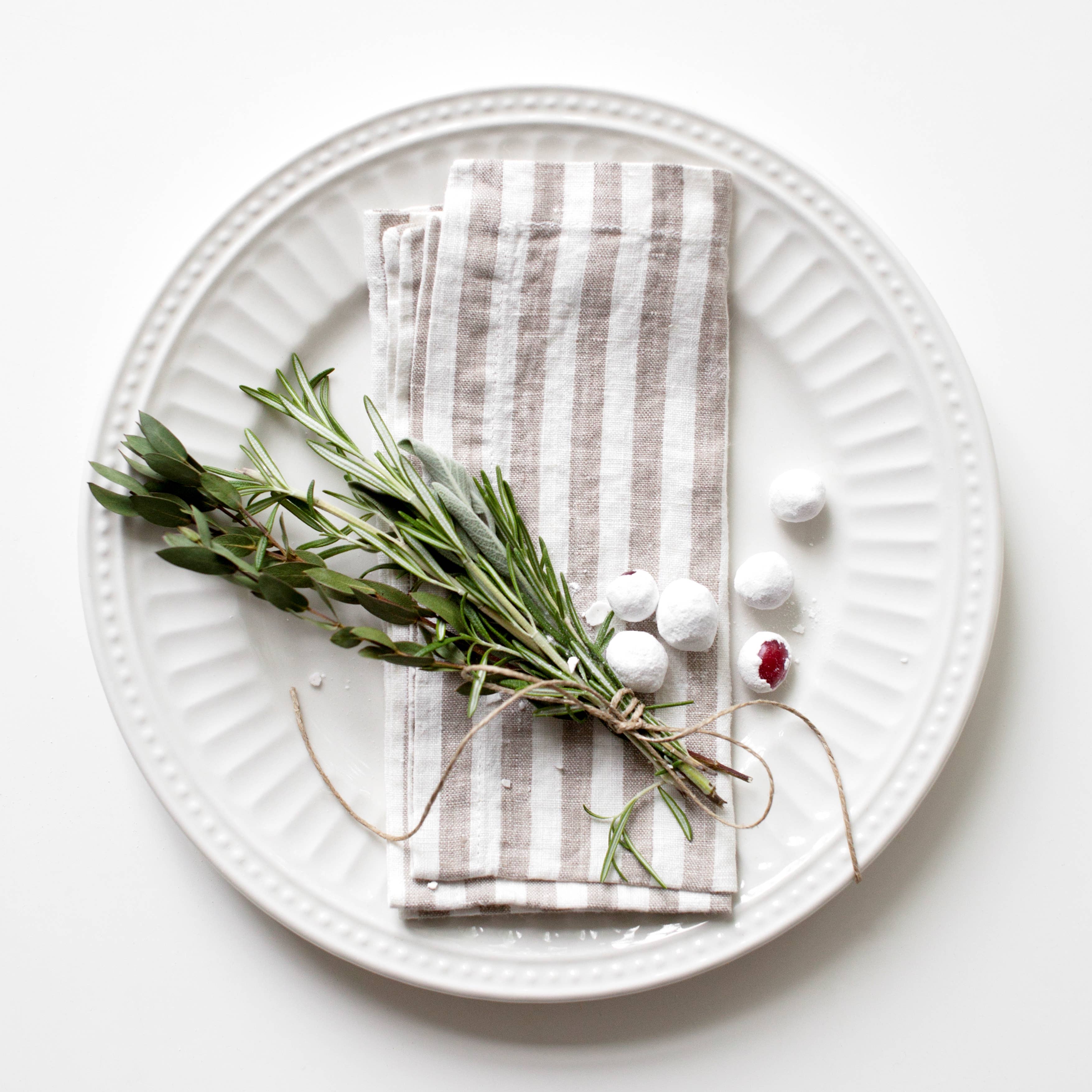 White plate with a white and natural striped linen napkin, greenery, and white balls on a white background
