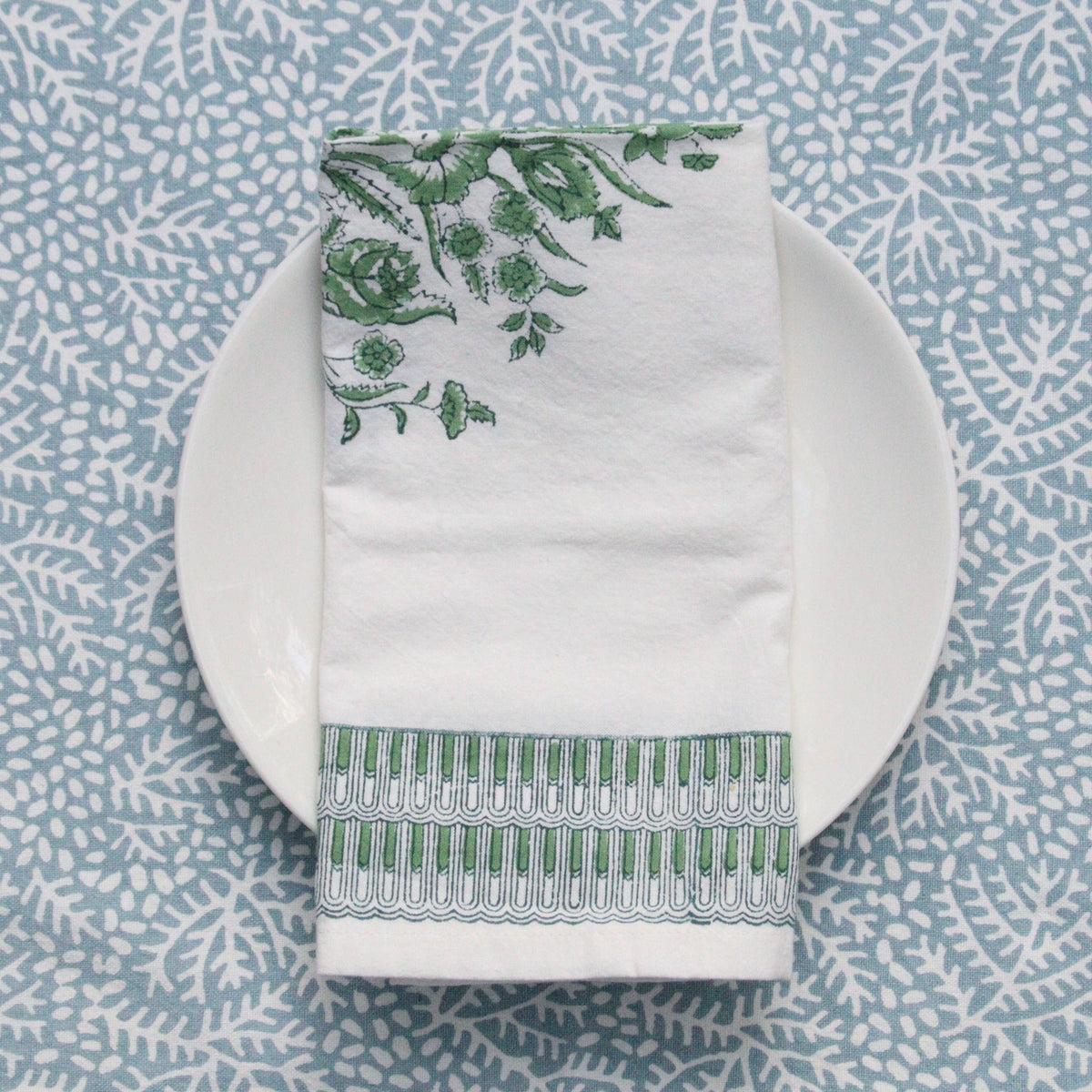 White napkin with green floral block print patterns on a white plate against a blue patterned background