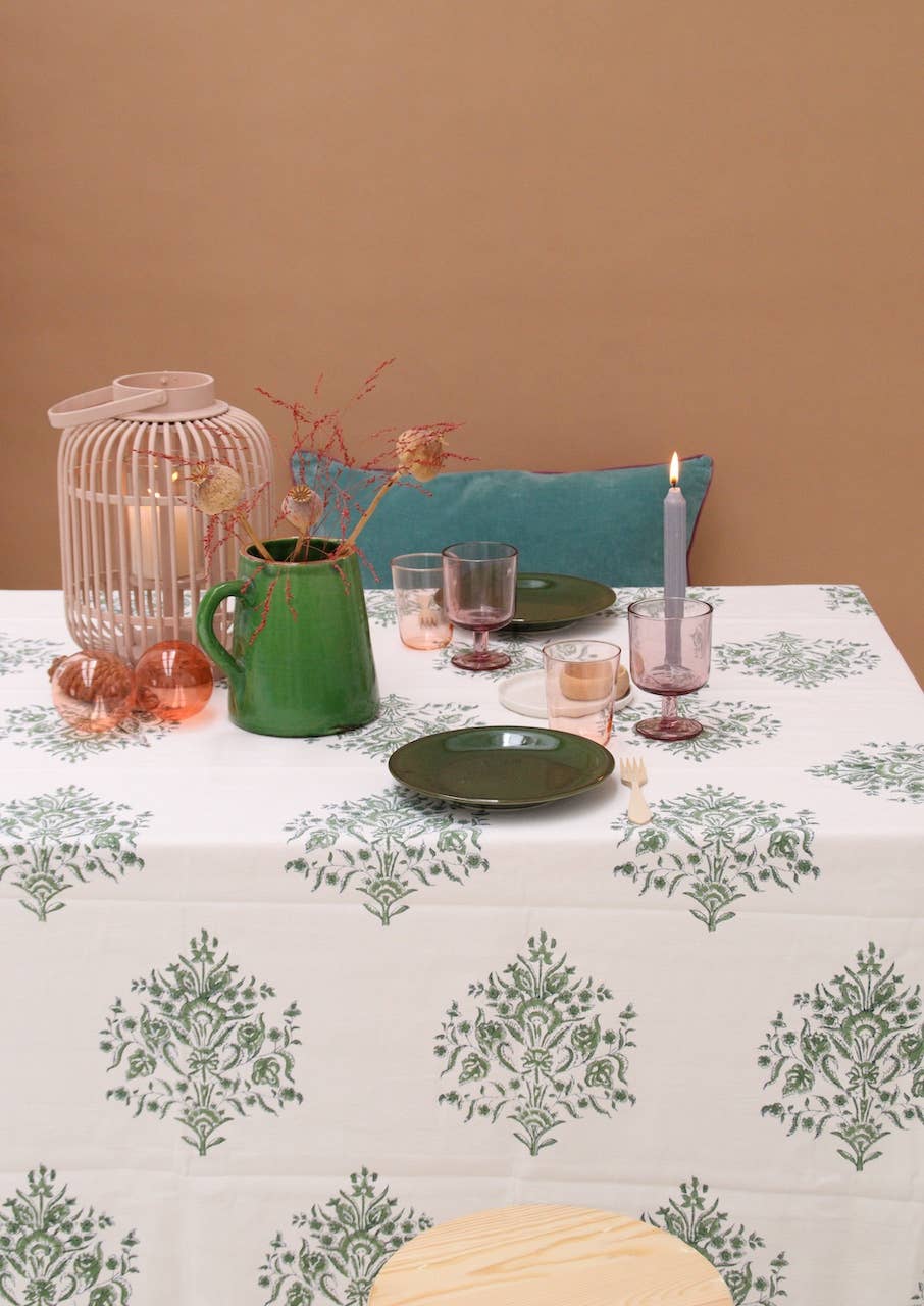 Dining table setting with a green, blue & white floral block print tablecloth, green pitcher, and pink candle against a brown wall.