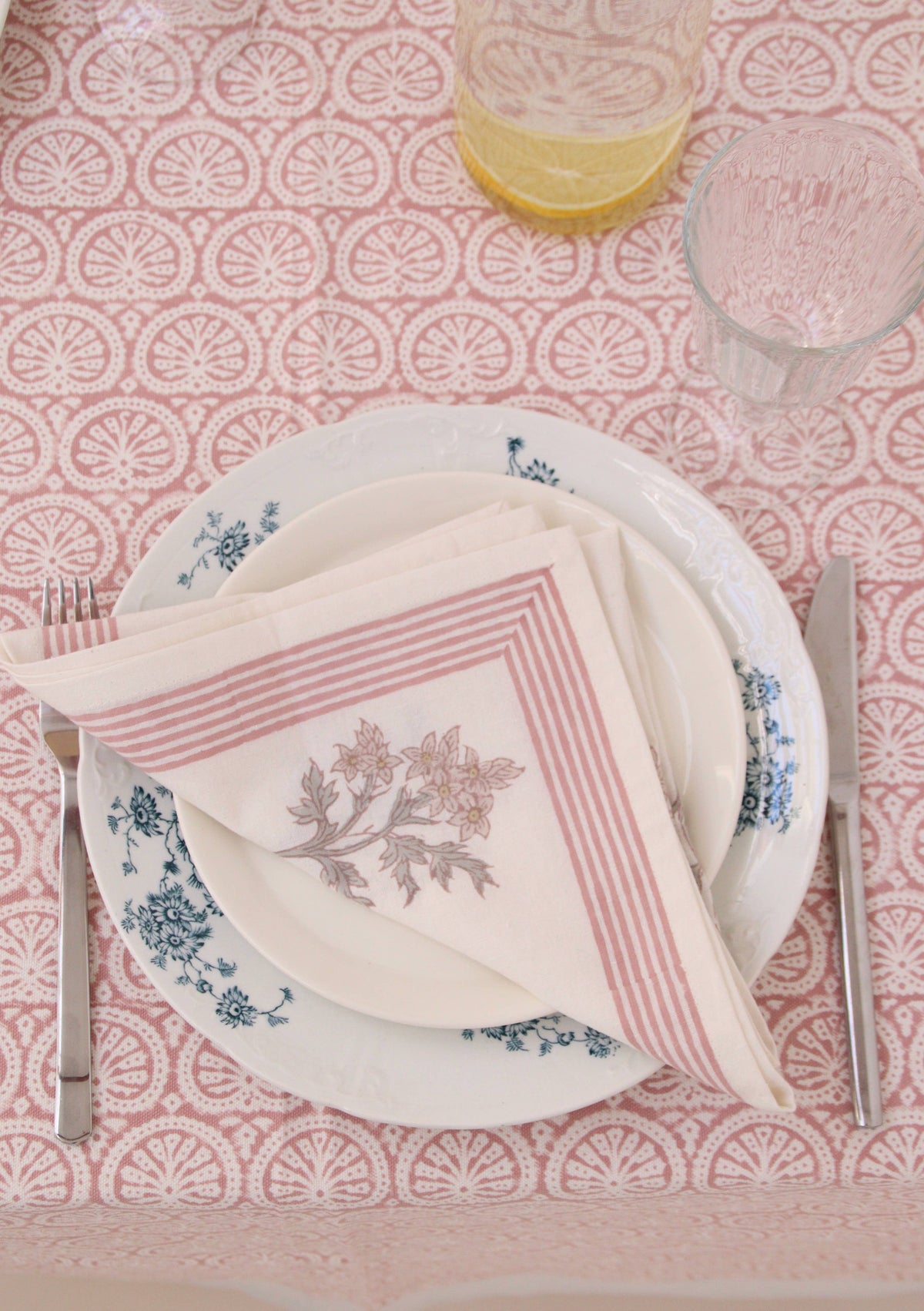 Table setting with floral plates, pink and white block print cotton napkins, and cutlery on a pink patterned tablecloth.