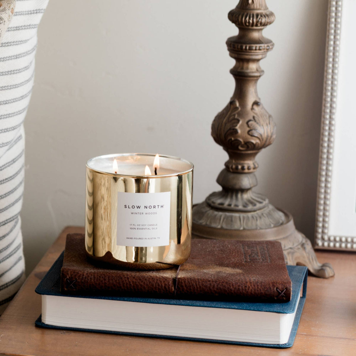A gold metal scented candle jar with a white label reading 'SLOW NORTH WINTER WOODS' sits on stacked books on a wooden table, with a large ornate lamp base and part of a striped cushion in the background.