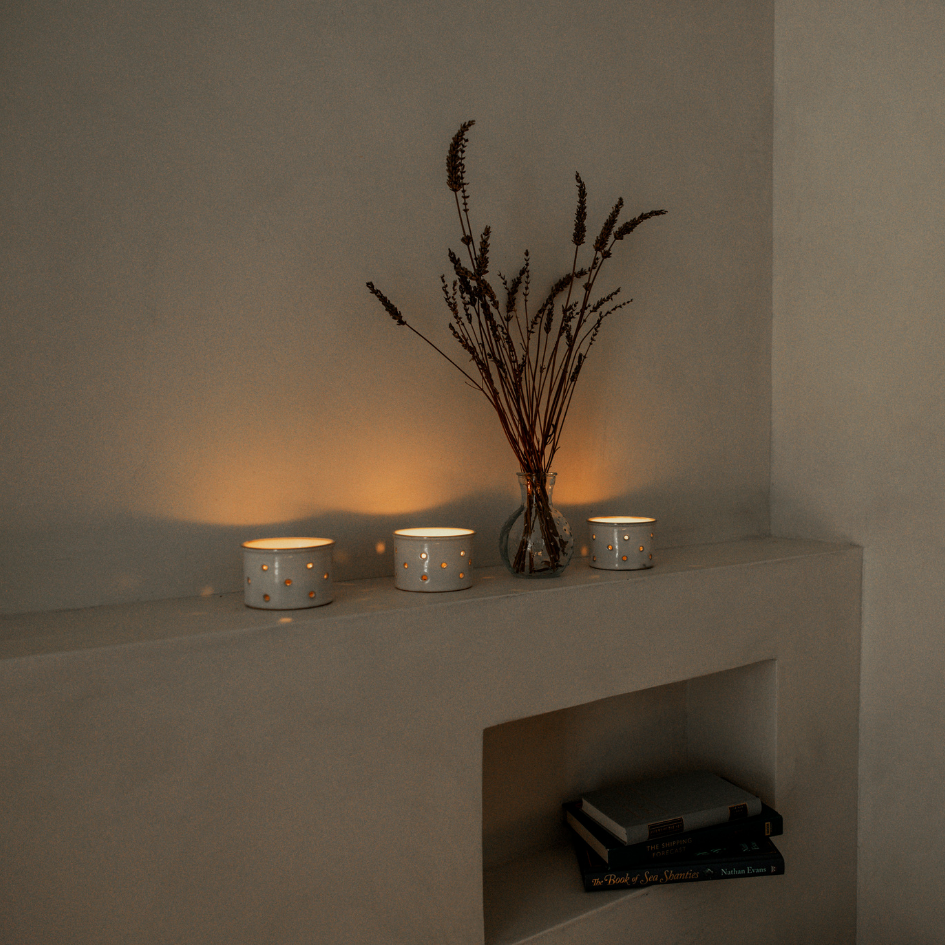Three small St Eval scented tea light candles and a vase with dried flowers on a concrete surface against a plain wall.