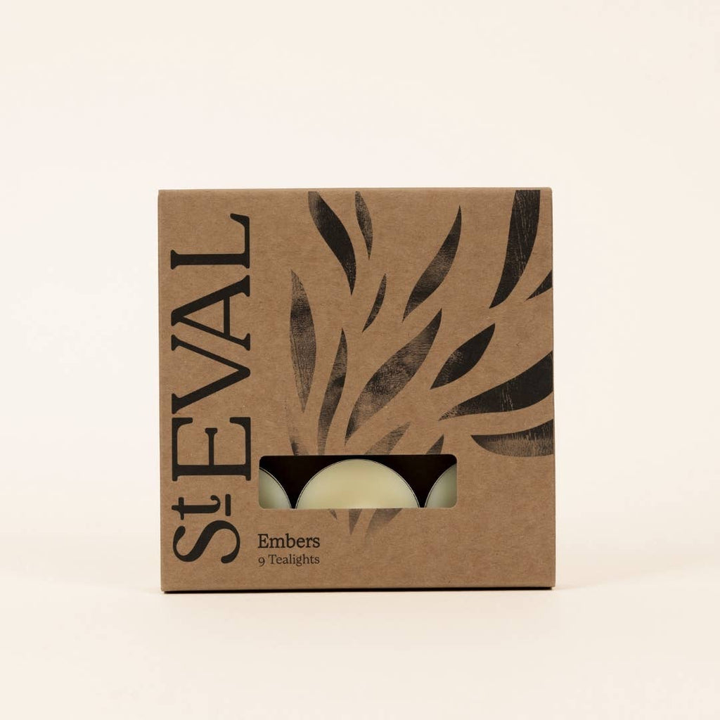 A square kraft paper box for St Eval Embers 9 scented tea lights, featuring a window that reveals two tea lights inside. The box is decorated with black leaf cutouts and bold vertical 'St EVAL' lettering