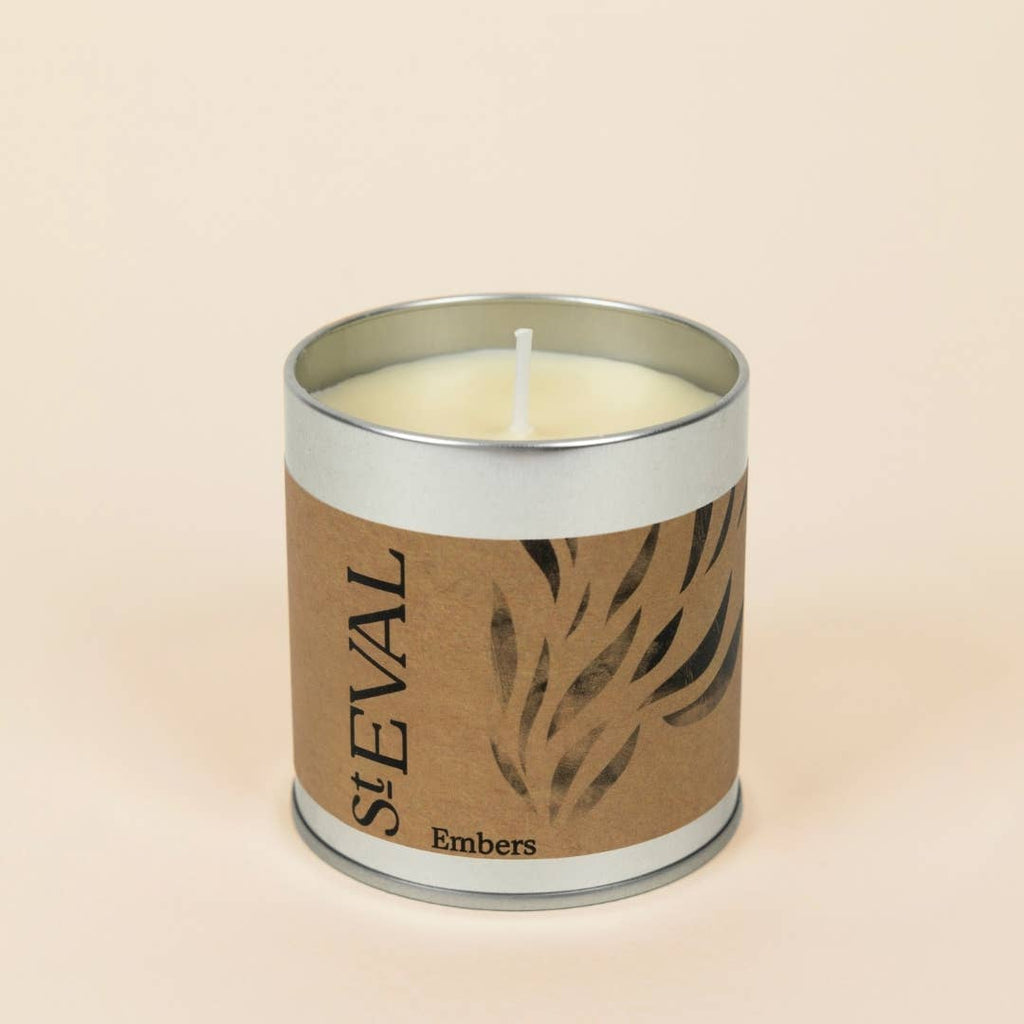 A single-wick scented candle in a cylindrical silver metal tin with a brown kraft paper label reading 'StEVAL Embers' and a leaf pattern, set against a solid light beige background.
