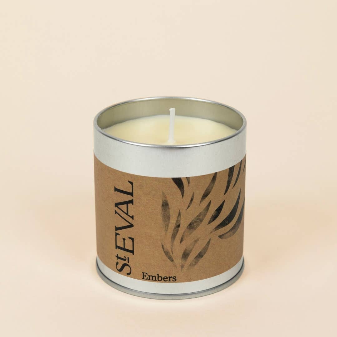 A single-wick scented candle in a cylindrical silver metal tin with a brown kraft paper label reading 'StEVAL Embers' and a leaf pattern, set against a solid light beige background.