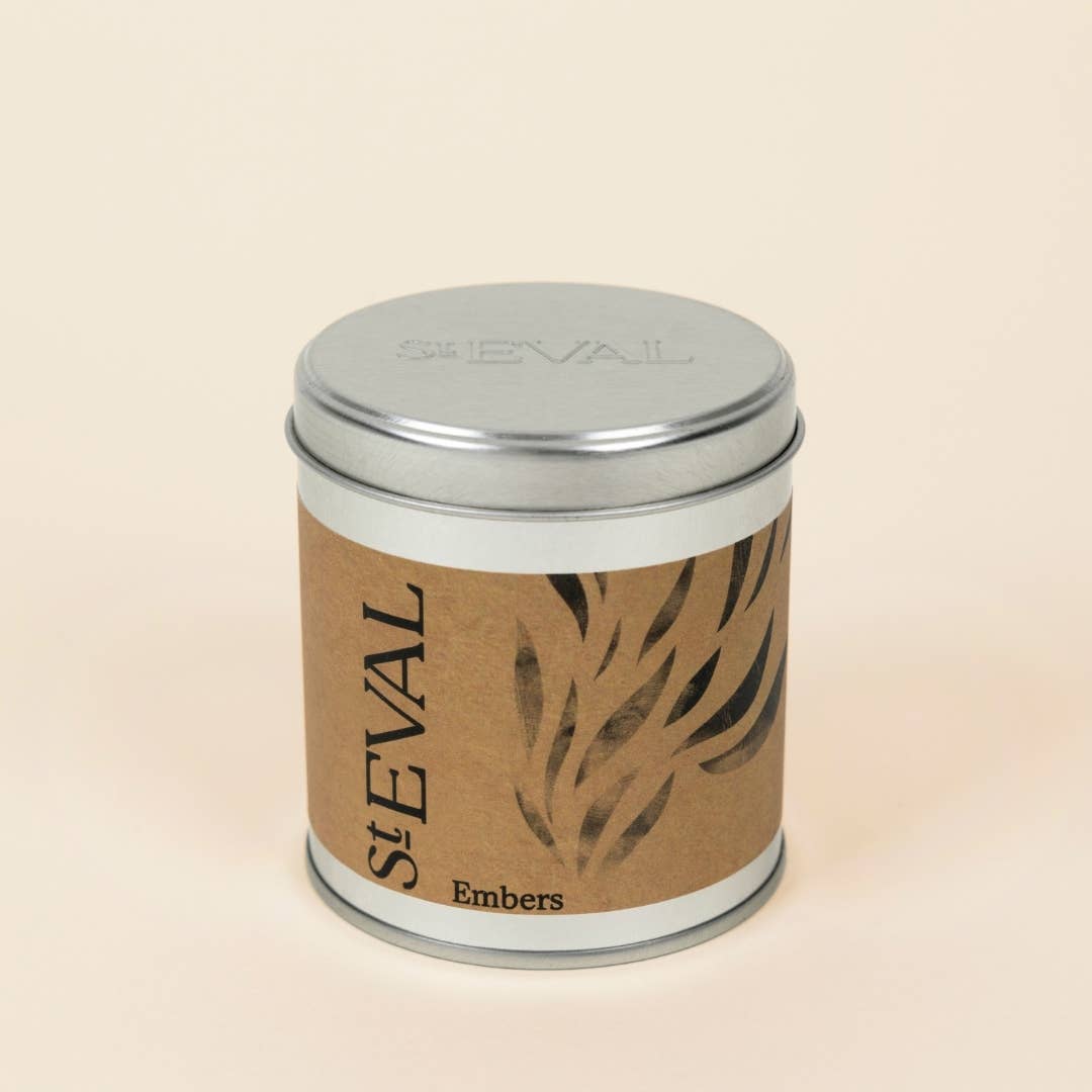 A cylindrical metal tin scented candle with a silver lid and a brown kraft paper label featuring the brand name 'St EVAL' and the scent 'Embers', set against a solid light beige background.
