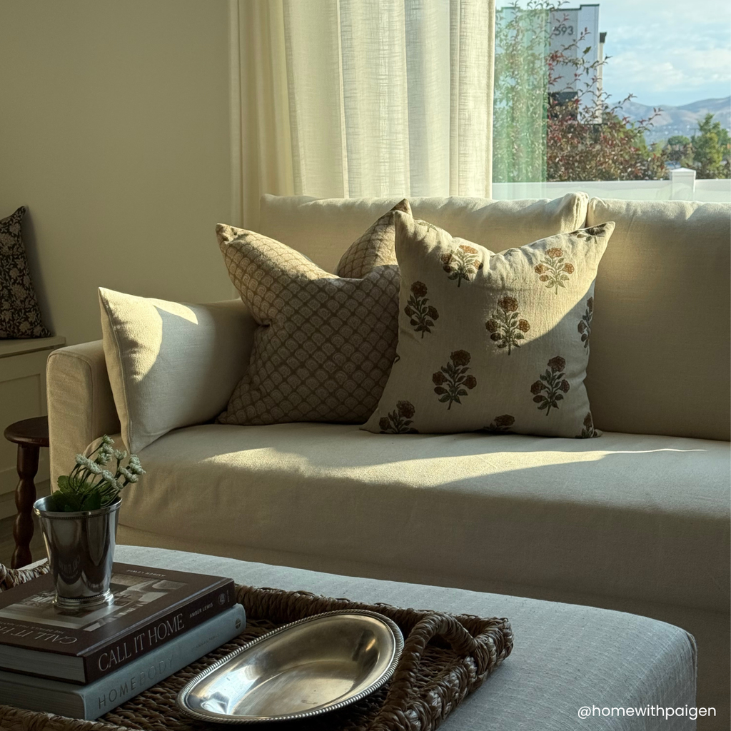 A styled living room scene with a cream sofa adorned with three decorative throw pillows in neutral tones, with a coffee table and books in the foreground.